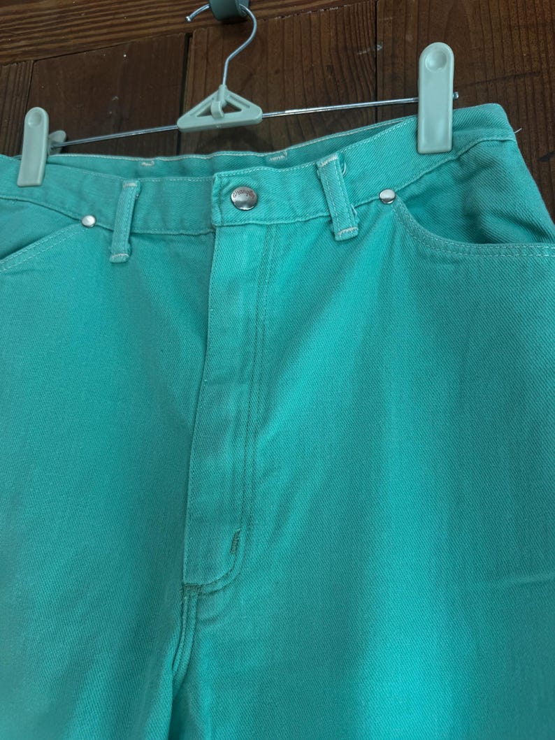 May include: Close-up of turquoise denim jeans. The jeans have a button closure, belt loops, and pockets. The jeans are hanging on a white and gray hanger against a brown wooden background. The jeans are a solid color.