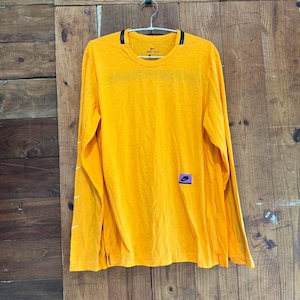 Puede incluir: Una camiseta de manga larga color amarillo mostaza con cuello redondo, que presenta un peque&ntilde;o logotipo Nike negro en el pecho, un parche rectangular morado con un swoosh negro en la parte inferior y logos blancos en la manga derecha.