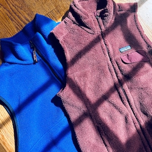 Puede incluir: Un chaleco de forro polar azul brillante de L.L.Bean con cremallera completa y un chaleco de forro polar granate oscuro de Patagonia con cuello alto, cremallera completa y un peque&ntilde;o bolsillo en el pecho con solapa. Ambos son sin mangas.