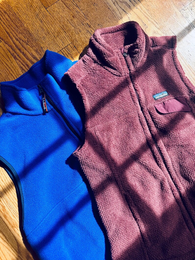 Puede incluir: Un chaleco de forro polar azul brillante de L.L.Bean con cremallera completa y un chaleco de forro polar granate oscuro de Patagonia con cuello alto, cremallera completa y un peque&ntilde;o bolsillo en el pecho con solapa. Ambos son sin mangas.