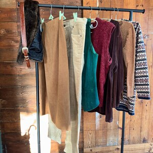 May include: A clothing rack displays a variety of apparel. Items include a brown faux fur vest, a brown leather belt, a tan skirt, beige pants, a green tank top, a burgundy sweater vest, a brown long-sleeve shirt, and a patterned sweater.