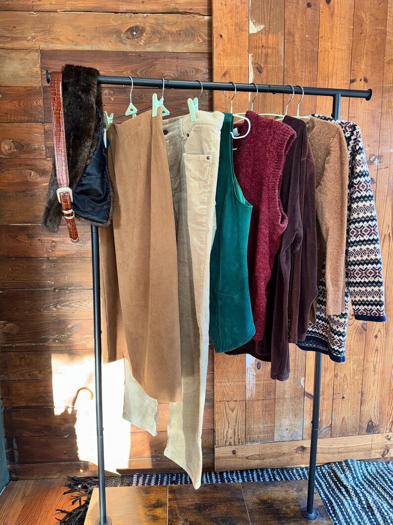 May include: A clothing rack displays a variety of apparel. Items include a brown faux fur vest, a brown leather belt, a tan skirt, beige pants, a green tank top, a burgundy sweater vest, a brown long-sleeve shirt, and a patterned sweater.