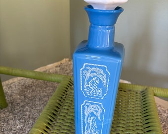May include: A blue glass decanter with a white ceramic stopper. The decanter has a square body with two white panels featuring a pastoral scene with a man and a dog.