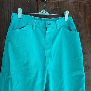 May include: A pair of turquoise denim pants hanging on a white hanger. The pants have a high waist, front pockets, and a button closure. The denim is a solid, vibrant turquoise color.