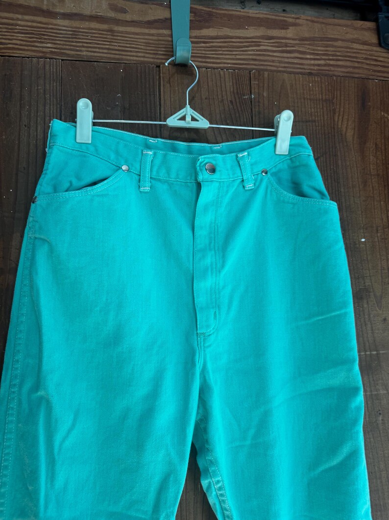 May include: A pair of turquoise denim pants hanging on a white hanger. The pants have a high waist, front pockets, and a button closure. The denim is a solid, vibrant turquoise color.