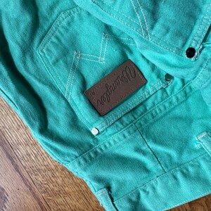 May include: Close-up of turquoise denim fabric with white stitching. A brown leather patch with the "Wrangler" logo is visible. The denim has a classic pocket design and metal rivets.