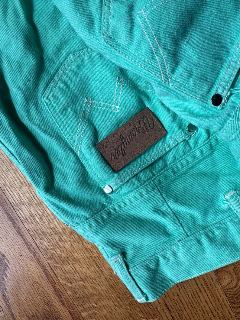 May include: Close-up of turquoise denim fabric with white stitching. A brown leather patch with the "Wrangler" logo is visible. The denim has a classic pocket design and metal rivets.