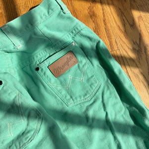 May include: Close-up of mint green Wrangler jeans. The jeans feature a brown leather patch with the Wrangler logo on the back pocket. White stitching accents the pockets and seams. The jeans are made of a sturdy denim material.