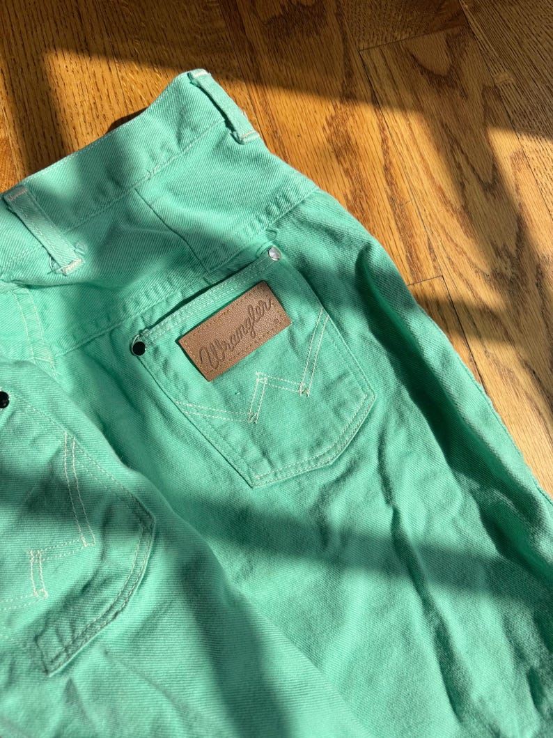 May include: Close-up of mint green Wrangler jeans. The jeans feature a brown leather patch with the Wrangler logo on the back pocket. White stitching accents the pockets and seams. The jeans are made of a sturdy denim material.