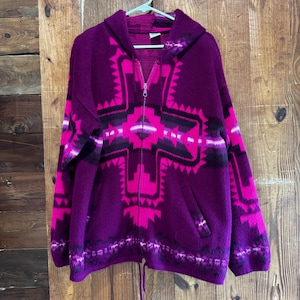 May include: A hooded zip-up sweater in shades of purple and pink, featuring a geometric pattern across the front and sleeves. The sweater has a drawstring at the waist and is hanging on a white hanger against a wooden background.
