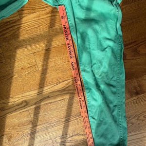 May include: Green denim pants with a straight leg design. A measuring tape shows the length of the pants. The text on the tape reads "HUNTER SAVINGS ASSN." and "Eight Convenient Locations."
