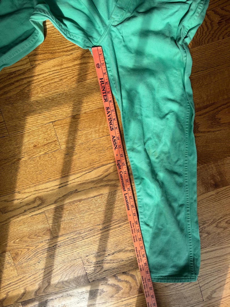 May include: Green denim pants with a straight leg design. A measuring tape shows the length of the pants. The text on the tape reads "HUNTER SAVINGS ASSN." and "Eight Convenient Locations."