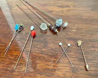 May include: A collection of vintage hat pins with various colored glass and metal embellishments. The pins are arranged on a wooden surface.