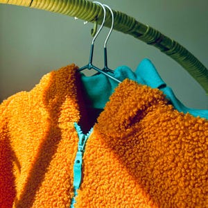 Puede incluir: Una chaqueta de forro polar sherpa de color naranja brillante con un exterior texturizado y difuso y una cremallera de color azul cerceta en contraste. La chaqueta cuelga de una percha de metal oscuro, mostrando un forro interior azul.