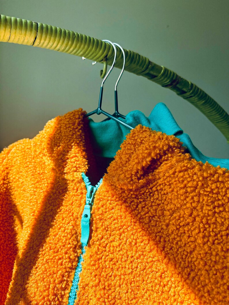 Puede incluir: Una chaqueta de forro polar sherpa de color naranja brillante con un exterior texturizado y difuso y una cremallera de color azul cerceta en contraste. La chaqueta cuelga de una percha de metal oscuro, mostrando un forro interior azul.