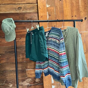 Op de afbeelding: Een groene baseballpet, groene shorts, een blauw-wit gestreepte trui en een groen lang mouw shirt hangen aan een zwarte rek tegen een houten muur.