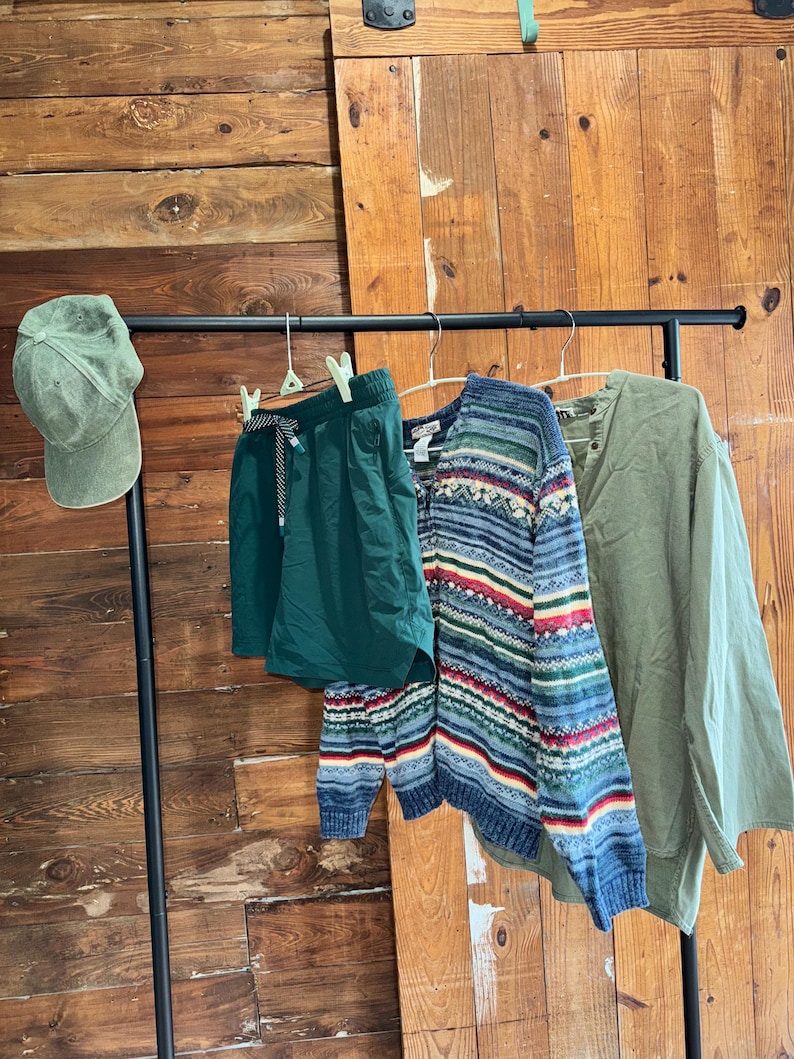Op de afbeelding: Een groene baseballpet, groene shorts, een blauw-wit gestreepte trui en een groen lang mouw shirt hangen aan een zwarte rek tegen een houten muur.