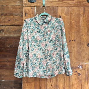 May include: A vintage floral print blouse with long sleeves and a collared neckline. The blouse features a pattern of pink and teal roses on a beige background. It is hanging on a green hanger against a wooden door.