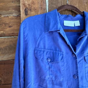 May include: A vibrant blue button-down shirt with two front pockets and long sleeves. The shirt is hanging on a wooden hanger against a wooden background. The shirt has a collared neckline and button closures.