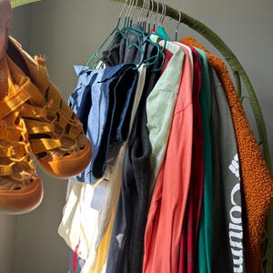 Puede incluir: Un par de sandalias cerradas de color amarillo mostaza sostenidas junto a un perchero curvo verde que exhibe varias prendas, incluyendo una camisa azul, un top blanco, un chaleco negro, una camisa coral de manga larga, una camisa roja estampada y una chaqueta Columbia verde oscuro.