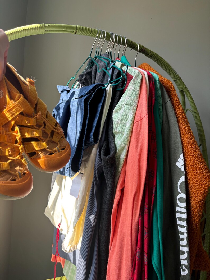 Puede incluir: Un par de sandalias cerradas de color amarillo mostaza sostenidas junto a un perchero curvo verde que exhibe varias prendas, incluyendo una camisa azul, un top blanco, un chaleco negro, una camisa coral de manga larga, una camisa roja estampada y una chaqueta Columbia verde oscuro.