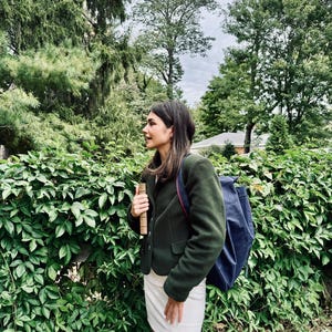 May include: A dark green blazer, white skirt, and navy blue backpack. The person is holding a book. The background includes a green hedge and trees. The image is taken outdoors on a cloudy day.