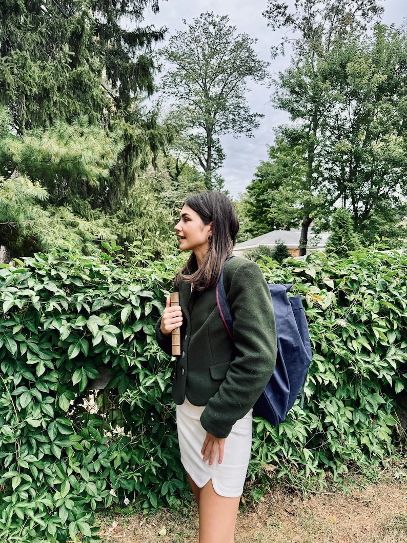 May include: A dark green blazer, white skirt, and navy blue backpack. The person is holding a book. The background includes a green hedge and trees. The image is taken outdoors on a cloudy day.