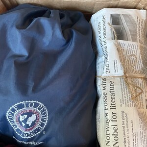 May include: A navy blue drawstring bag with a white and purple logo, and a bundle of newspapers tied with twine inside a cardboard box. The newspaper headlines include "Norway's Fosse wins Nobel for literature."