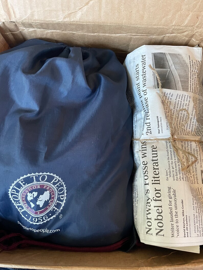 May include: A navy blue drawstring bag with a white and purple logo, and a bundle of newspapers tied with twine inside a cardboard box. The newspaper headlines include "Norway's Fosse wins Nobel for literature."