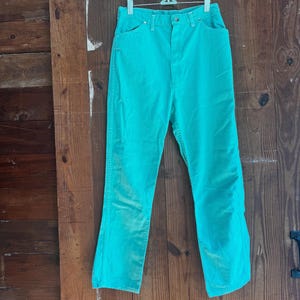 May include: A pair of turquoise denim jeans hangs from a metal hanger against a wooden door. The jeans have a classic five-pocket design, a button and zipper closure, and a high waist. The fabric appears slightly faded.