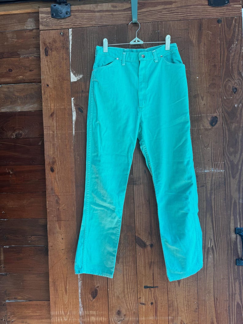 May include: A pair of turquoise denim jeans hangs from a metal hanger against a wooden door. The jeans have a classic five-pocket design, a button and zipper closure, and a high waist. The fabric appears slightly faded.