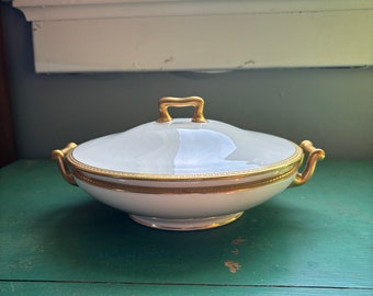 May include: A white porcelain serving dish with a lid. The dish has gold trim around the rim and handles. The lid has a gold handle. The dish is sitting on a green surface.