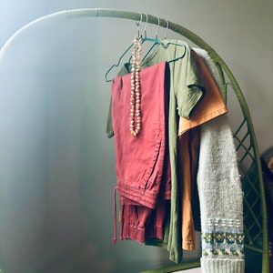 Op de afbeelding: Een groene rieten kledingrek met verschillende kledingstukken die eraan hangen. De kleding omvat een ​​rode broek, een groen T-shirt, een beige T-shirt en een grijze trui. Een kralenketting hangt aan het rek.