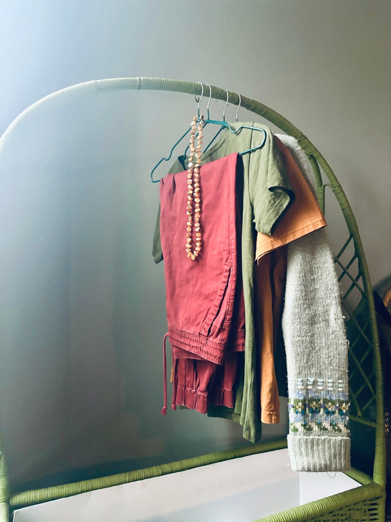 Op de afbeelding: Een groene rieten kledingrek met verschillende kledingstukken die eraan hangen. De kleding omvat een ​​rode broek, een groen T-shirt, een beige T-shirt en een grijze trui. Een kralenketting hangt aan het rek.