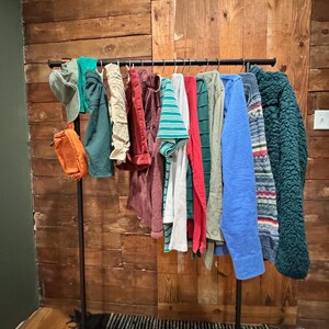 Op de afbeelding: Een zwarte metalen kledingrek met verschillende kledingstukken die eraan hangen. De kleding omvat een groen-wit gestreept shirt, een rood shirt, een groene trui, een blauwe trui, een groen-wit gepatroneerde trui en een bruine trui.