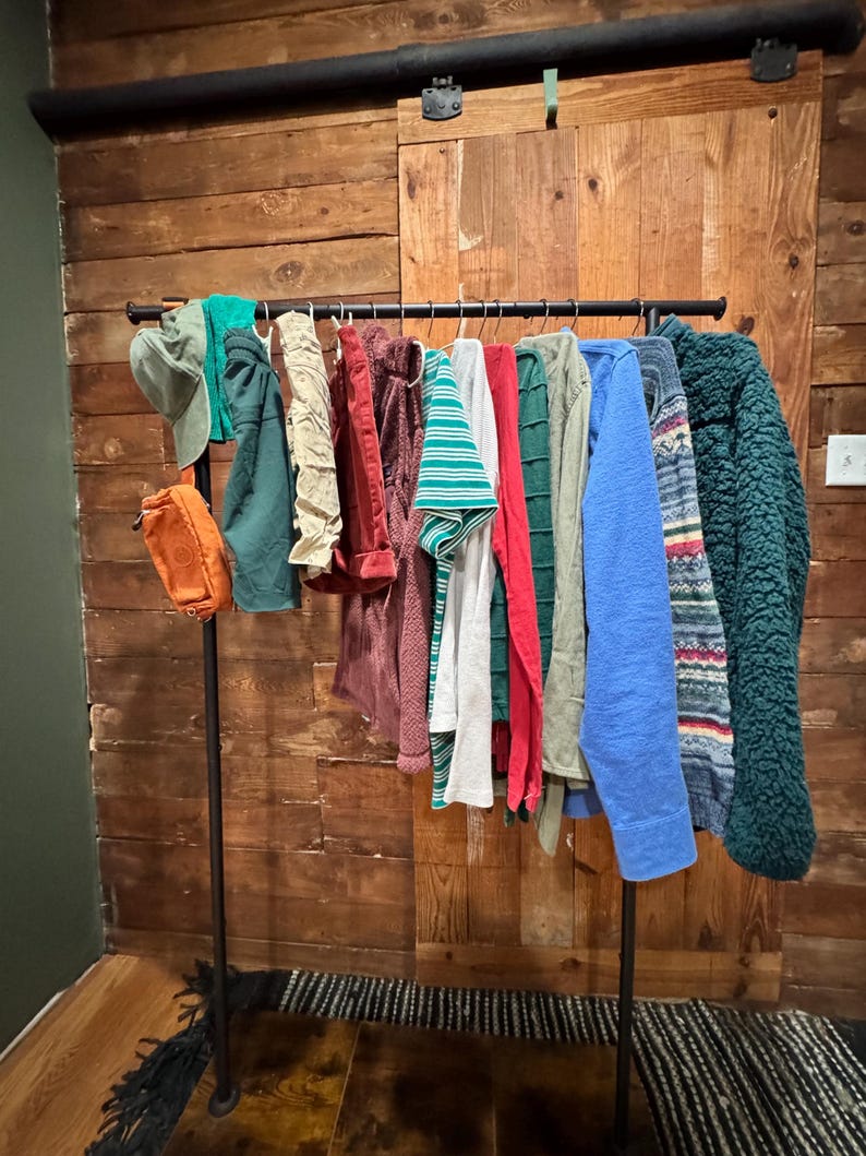 Op de afbeelding: Een zwarte metalen kledingrek met verschillende kledingstukken die eraan hangen. De kleding omvat een groen-wit gestreept shirt, een rood shirt, een groene trui, een blauwe trui, een groen-wit gepatroneerde trui en een bruine trui.