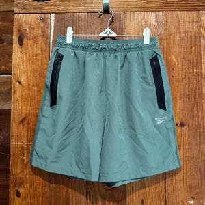 Puede incluir: Pantalones cortos deportivos de color verde salvia con cintura el&aacute;stica, dos bolsillos laterales con cremallera negra y el logotipo de Reebok en blanco impreso en la pierna derecha. Est&aacute;n hechos de tela lisa y ligera con corte holgado.