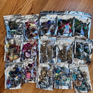 May include: Assortment of 15 clear plastic bags filled with various colorful jewelry findings, including beads, charms, and metal pieces. The bags are arranged in three rows of five on a wooden surface.