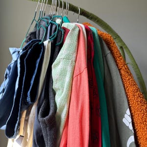 Puede incluir: Una colecci&oacute;n de varias prendas colgadas en un perchero verde curvado, incluyendo un pantal&oacute;n azul, una camisa blanca, un chaleco gris oscuro, una blusa verde claro texturizada, una camisa coral de manga larga, una camisa roja estampada y una prenda de forro polar naranja.