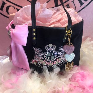 May include: A black tote bag featuring a large embroidered crest with a crown, two dogs, and the text JUICY COUTURE. The bag is adorned with a large pink satin bow, gold-tone hardware, and a dangling charm set with two heart-shaped crystals.