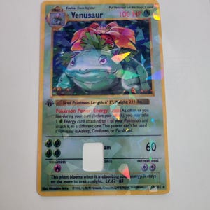 Puede incluir: Una carta hologr&aacute;fica de Venusaur Pok&eacute;mon. La carta presenta un Venusaur verde y azul con una gran flor roja y verde en su espalda. La carta tiene el texto "Venusaur" y "100 HP".