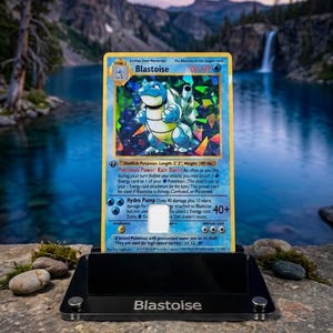 May include: A holographic Blastoise Pokémon card, displayed in a black stand, with a scenic background of a lake and mountains. The card features a blue and white turtle-like creature with the text "Blastoise" and "100 HP".