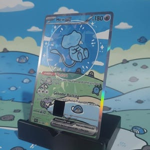 May include: A holographic Mew ex Pokémon trading card, displayed in a black stand. The card features a light blue Mew character within a blue circle, with text including "Ability Restart" and "Genome Hacking".