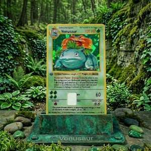 May include: A Venusaur Pokémon trading card, featuring a green and orange creature, displayed on a bronze-colored stand. The card has text including "100 HP" and "Venusaur". The background is a lush forest setting.
