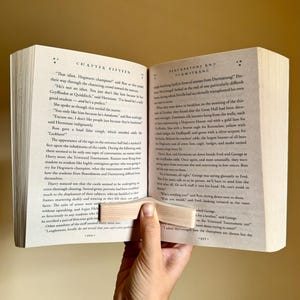May include: A close-up of an open book with a wooden page holder. The book is titled "Harry Potter and the Goblet of Fire" and the page is open to Chapter Fifteen, "Beauxbatons and Durmstrang". The text on the page discusses the Triwizard Tournament and the upcoming competition between Hogwarts, Beauxbatons, and Durmstrang.