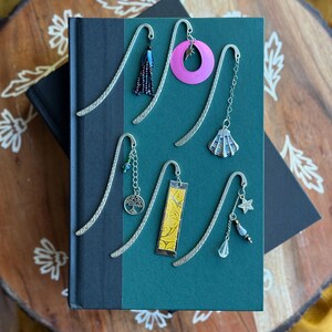 May include: A collection of decorative metal bookmarks with various charms, including a tassel, a pink circle, a seashell, a tree of life, a yellow rectangle, and a star. The bookmarks are placed on a closed book with a green cover.