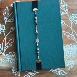 May include: A teal-colored journal with a black elastic band bookmark. The bookmark features gold-toned metal accents, pink and black beads, and a decorative arrow. The journal is on a wooden surface with floral designs.
