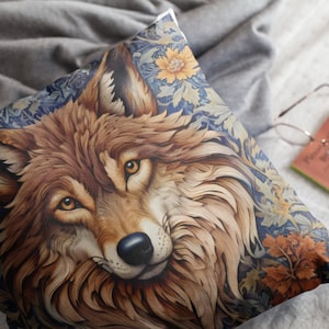 以下が含まれることがあります： オオカミの顔の精巧なイラストが描かれた正方形のクッション。オオカミは茶色の毛皮と金色の目を持ち、黄色とオレンジのアクセントが付いた青色の花柄の背景に配置されています。クッションは灰色の表面に置かれています。