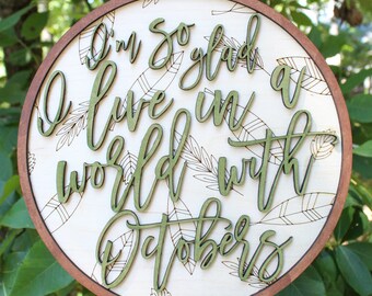 May include: A round wooden sign with a brown frame and a white background. The sign has a green leafy pattern and the words "I'm so glad I live in a world with Octobers" written in green cursive.