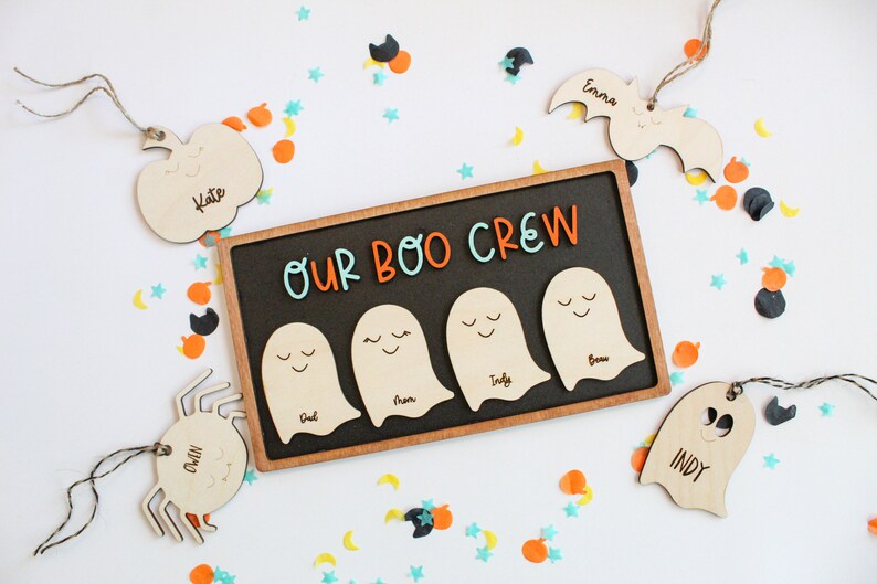 May include: A wooden sign with the text "Our Boo Crew" in orange and blue letters. Four wooden ghost shapes are attached to the sign, each with a different name written on them. The sign is surrounded by Halloween-themed confetti.
